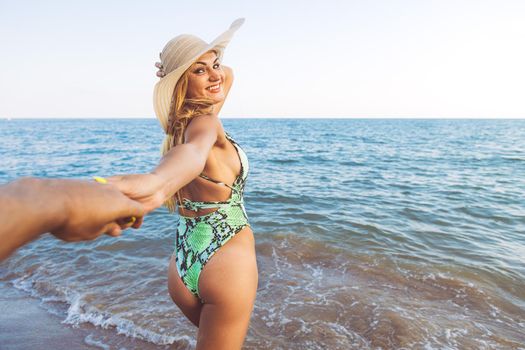follow me shot of a smiling blonde woman with straw hat and swimsuit leading her boyfriend on sea shore, concept of honeymoon and summer vacation