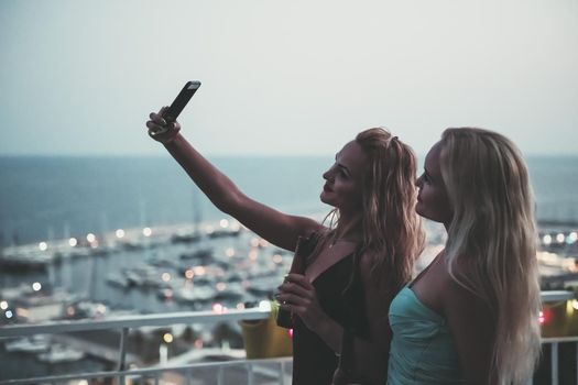 two beautiful young girls with beers taking a photo with the phone in a private party on the outdoor terrace at the night, leisure happiness and friendship concept, vintage look with grain