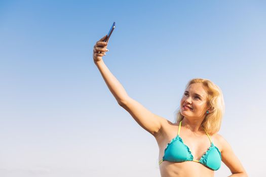 blonde woman in swimsuit making a video call with her mobile phone with a clear blue sky in the background on her summer vacation, communication and technology concept