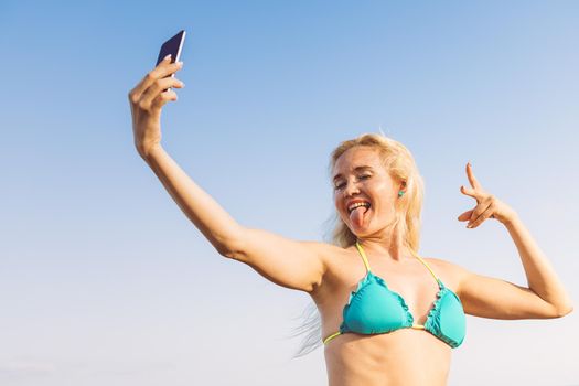 blonde woman in swimsuit taking a selfie photo with her mobile phone with a clear blue sky in the background on her summer vacation, communication and technology concept