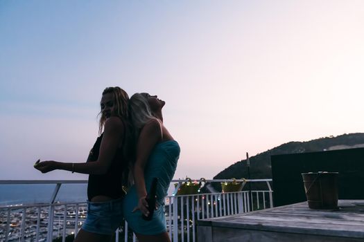 silhouette of two beautiful young girls with beers dancing and having fun at a private party on the outdoor terrace at the night, leisure happiness and friendship concept, vintage look with grain