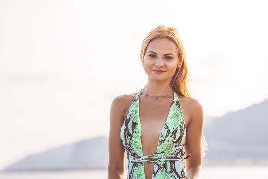 sensual blonde woman in a sexy green swimsuit smiling at the sunset on the beach, concept of glamour and summer vacation, copy space for text