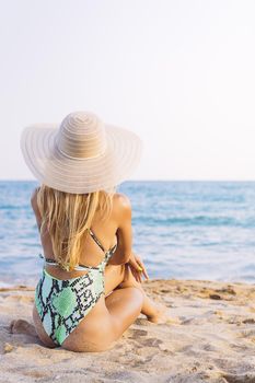 vertical photo of the back of an unrecognizable blonde woman with a hat and a sexy green swimsuit sitting on the beach, concept of glamour and summer vacation, copy space for text