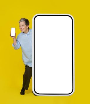 Looking out behind big smartphone mature woman with grey haired smiling holding smartphone showing it white screen. Pretty woman in blue blouse isolated on yellow background. Mobile app advertising.