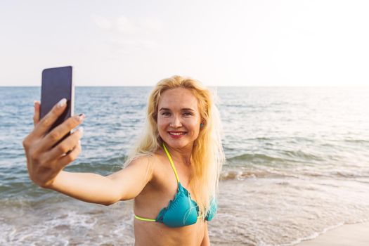 blonde woman making a video call with her mobile phone from the beach at sunset on her summer vacation, communication and technology concept