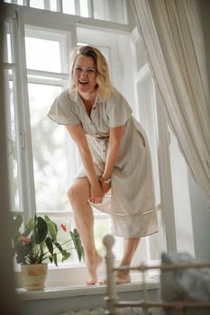 Lifestyle portrait of a young lady having fun in a sunlit room. Charming girl with blond hair, cream dress, standing on the windowsill, smiling and looking into the distance.