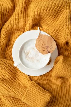 cup of cappuccino coffee with yellow leaf on yellow sweater top view flat lay