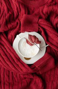 cup of cappuccino coffee with red leaf on red sweater top view flat lay