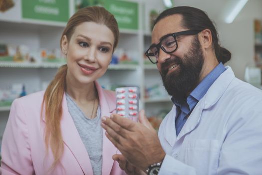 Mature experienced pharmacist smiling joyfully helping young woman choosing medication. Bearded chemist offering female customer pills, working at drugstore. Woman shopping for medicine