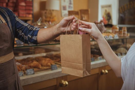 Cropped shot of a male baker wearing apron giving paper shopping bag to female customer, copy space. Woman buying food at te bakery store. Consumerism, organic food, health concept