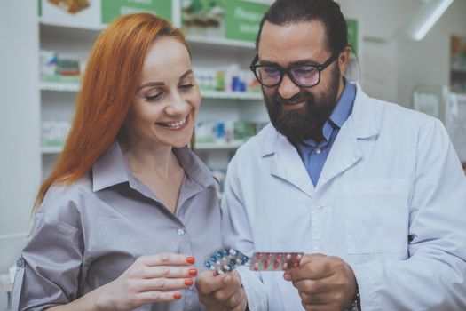 Beautiful mature red haired woman shopping for drugs at the pharmacy, examining pills professional druggist is offering her. Bearded mature pharmacist selling medications to a female customer