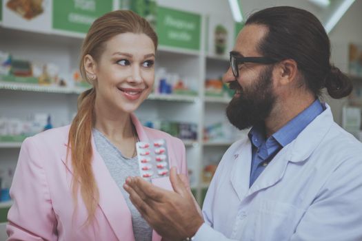 Lovely young woman smiling, buying pills at the drugstore. Bearded mature pharmacist helping his female customer showing her blister of pills. Female customer shopping for medicine at pharmacy