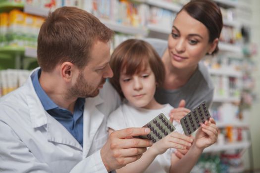 Mature male pharmacist helping choosing medicament for his female client and her little son. Beautiful young woman shopping at drugstore with her child