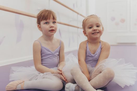Cute happy little girl wearing leotard and tutu skirs smiling to the camera sitting with her friend at ballet school. Adorable little ballerinas resting after exercising at dance school