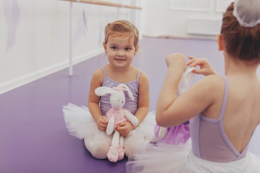 Cute happy little girl in leotard and tutu holding her toy, sitting with her best friend at ballet school, waiting for lesson. Little cute ballerinas relax before exercising at dance studio