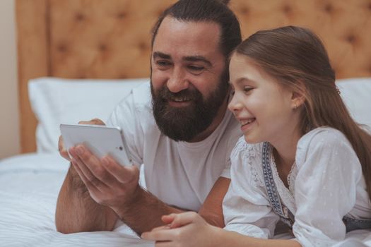 Close up of a handsome bearded mature man enjoying using smart phone with his adorable little daughter. Single father teaching his child how to use internet safely. Online, child safety concept