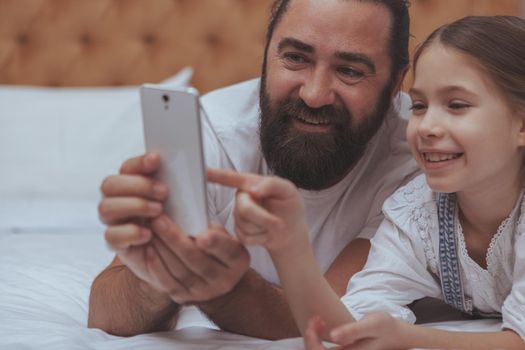 Cropped close up of a beautiful cheerful little girl browsing online, using smart phone with her loving father. Handsome bearded man smiling, enjoying spending weekend at home with his daughter, browsing online