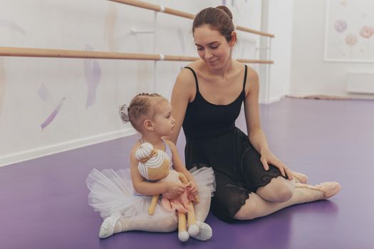 Beautiful young woman ballet dancer sitting relaxed with her little cute student, smiling at each other. Adorable little girl in tutu smiling at her dance teacher. Ballerina resting after dancing with little girl