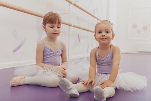 Two adorable cheerful little ballerinas rest on the floor at dance studio. Cute happy little girl in ballet outfit smiling joyfully, sitting with her friend after ballet class