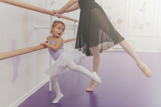 Graceful ballerina teaching ballet to adorable little girl in tutu and leotard. Cute little girl enjoying exercising at ballet school with her teacher, copy space. Guidance, growth, success concept