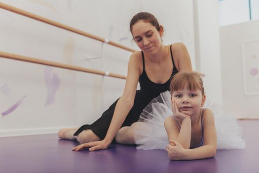 Lovely little ballerina looking to the camera, lying relaxed on the floor at ballet school after exercising with her teacher. Young beautiful woman enjoying teaching ballet to little girl, copy space