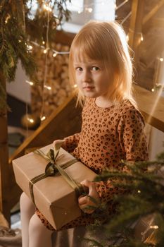 A little blonde girl is sitting on a wooden staircase in a Scandinavian interior decorated in a New Year's style. A child holds a Christmas gift in a craft package.