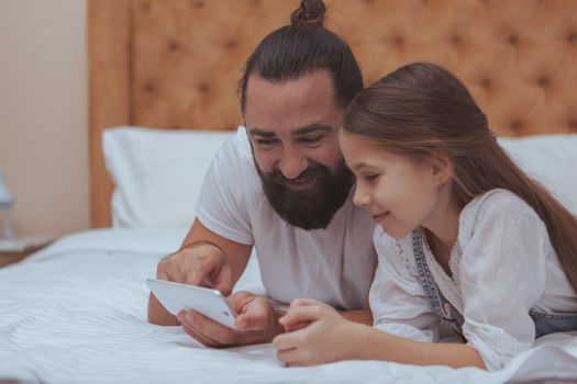 Cute happy little girl and her father using smart phone together, relaxing at home. Handsome bearded man showing his little daughter how to use mobile phone, copy space