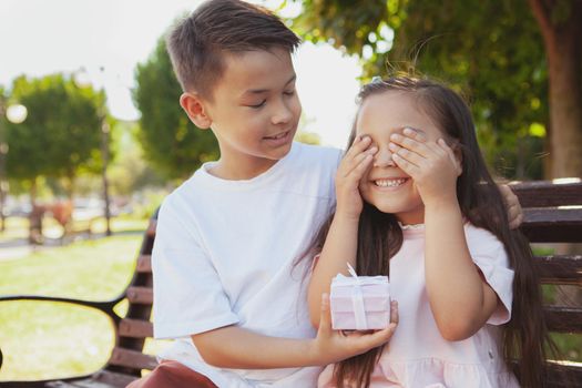 Cheerful Asian young boy giving a surprise gift to his little sister. Adorable little girl covering her eyes with her hands, receiving a present from her brother