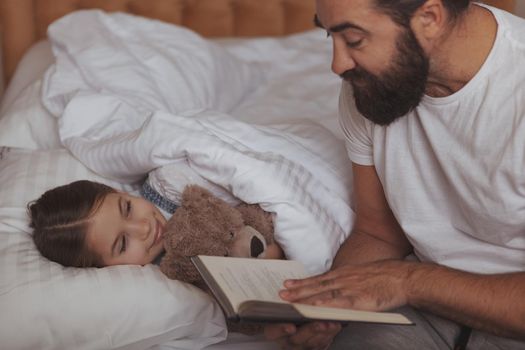 Loving father reading a fairytale to his adorable little daughter before sleep. Cheerful cute girl enjoying listening her dad read a book before night sleep