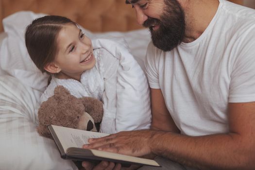 Adorable happy little girl holding teddy bear, smiling at her father, while he is reading her a fairytale good night. Cropped shot of a cheerful handsome bearded man getting his child to sleep, reading a book