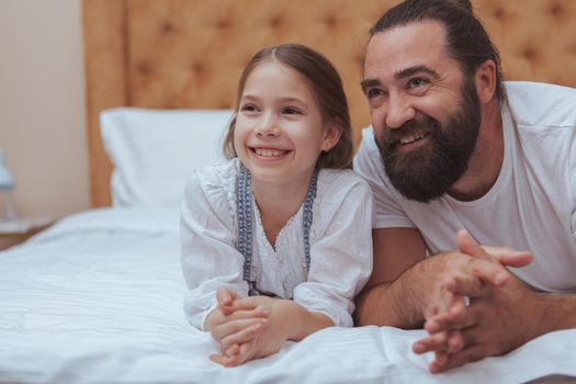 Handsome bearded mature man and his adorable little daughter lying together on bed, smiling looking away. Cheerful little girl and her dad relaxing at home together