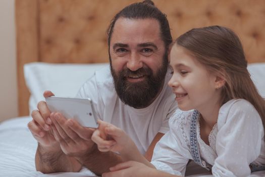 Handsome bearded man smiling to the camera, while using smart phone with his adorable daughter. Cute little girl smiling, using mobile phone with her dad. Online, internet, gadget concept