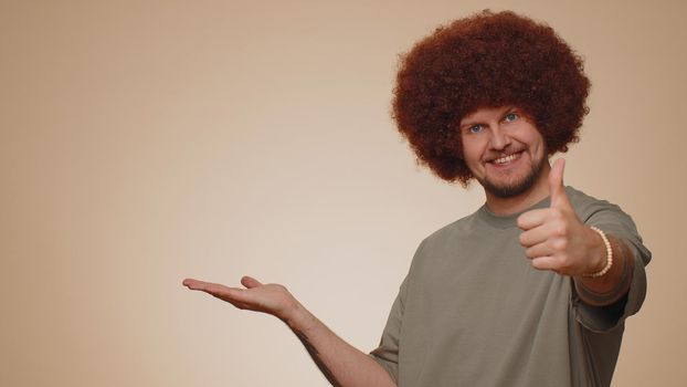 Handsome one bearded man showing thumbs up and pointing empty place, advertising area for commercial text, copy space for goods promotion. Young adult guy. Studio shot indoors on beige wall background