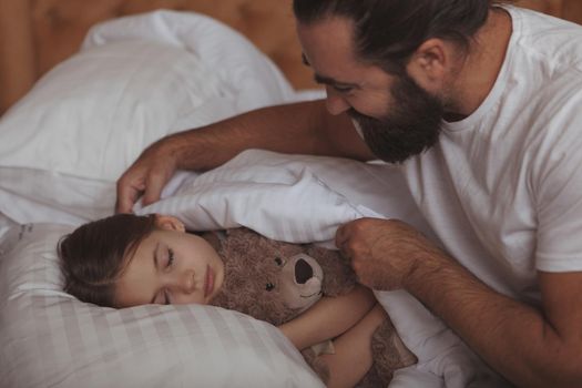 Loving father tucking blanket for his lovely little daughter. Adorable girl sleeping in bed, hugging her teddy bear, her father pulling up her blanket. Single parenting concept