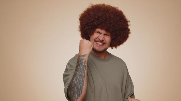 Happy joyful man in casual t-shirt shouting, raising fists in gesture I did it, celebrating success, winning, birthday, lottery goal achievemen. Adult guy boy indoors on beige studio wall background