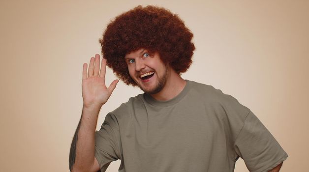Handsome bearded man in t-shirt smiling friendly at camera and waving hands gesturing hello or goodbye, welcoming with hospitable expression. Young happy guy indoors isolated on beige wall background
