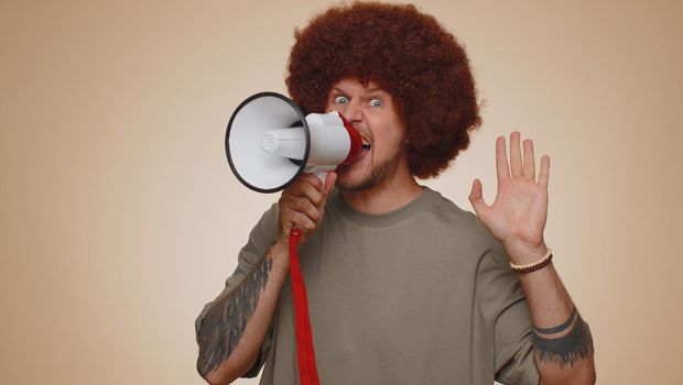 Smiling happy bearded man talking with megaphone, proclaiming news, loudly announcing sale advertisement, warning using loudspeaker to shout speech. Young handsome adult guy on beige studio background