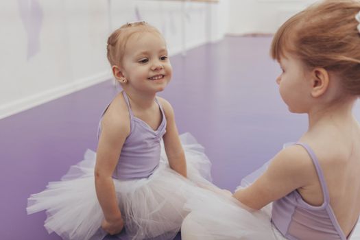 Adorable happy little girl smiling joyfully at her friend, resting together after ballet class at dance studio. Cute little ballerinas attending ballet school together. Communication, bonding concept