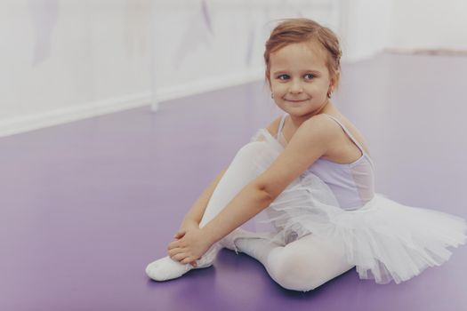 Cute little happy ballerina smiling to the camera sitting on the floor at ballet school. Adorable little girl in tutu and leotard relaxing after ballet lesson at dance studio, copy space