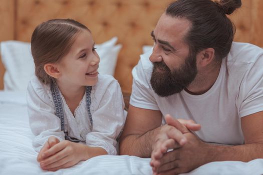 Handsome bearded man with manbun hairdo smiling at his adorable little daughter. Cute happy girl laughing joyfully, resting at home with her dad