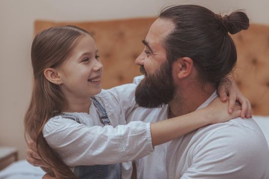 Happy father and daughter hugging, smiling at each other, having fun at home. Handsome bearded mature man embracing his adorable little daughter