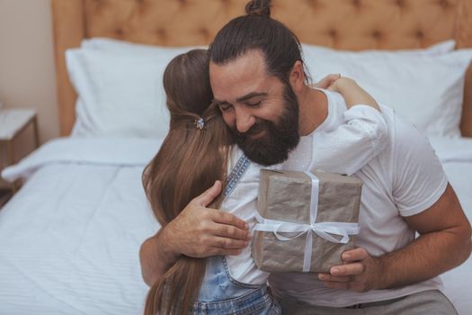 Happy handsome bearded man smiling, hugging his little daughter after receiving a present from her, copy space. Cute little girl giving a present to her dad. Father and daughter celebrating together