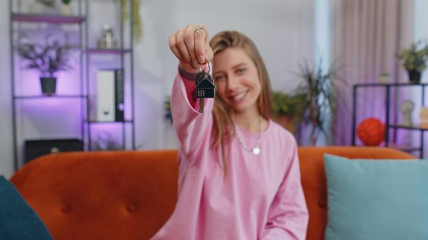 Young woman real estate agent lifting hand showing the keys of new home house apartment, buying or renting property, mortgage loan at modern home apartment indoors. Lovely girl in room sitting on sofa