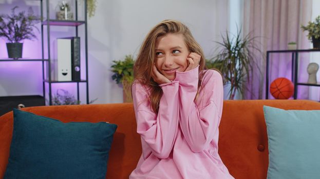 Close-up of happy calm girl in hoodie smiling friendly, glad expression looking away dreaming resting, relaxation feel satisfied concept good news. Young caucasian woman sitting on sofa at home room