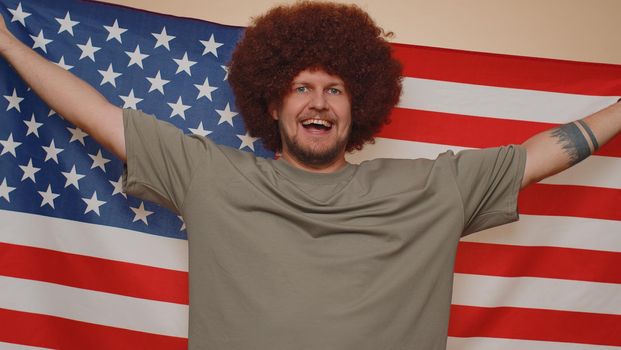 Handsome bearded man with lush hairstyle waving and wrapping in American USA flag, celebrating, human rights and freedoms. Independence day. Young adult guy boy isolated on beige studio background