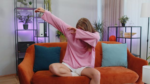 Trendy cheerful positive adult girl having fun dancing and moving to rhythm, dabbing raising hands, making dub dance gesture. Young caucasian woman at modern home apartment living room on sofa couch