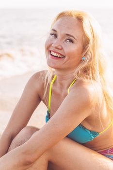 vertical portrait of a beautiful smiling blonde girl posing sitting on the beach sand, summer holiday concept and glamour