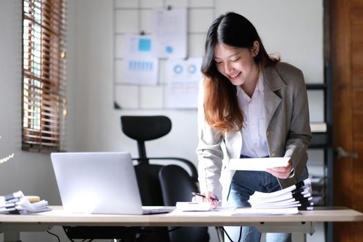 Beautiful Asian businesswoman standing and working on laptop with smiling face while working in office..