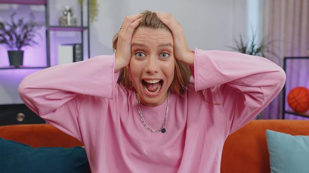 Oh my God, Wow. Lovely adult girl surprised looking at camera with big eyes, shocked by sudden victory, good win news, celebrating at home. Excited amazed young woman indoors in living room on couch