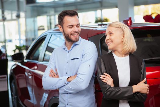 Lovely couple smiling at each other cheerfully, standing near their new car at the dealership. Husband and wife celebrating buying new automobile